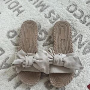 Beige sandaler med rosett - Snygga beige sandaler med flätad sula och breda band i mockaliknande material. Framtill pryds de av en stor rosett som ger en trendig och söt look. Perfekta för varma dagar och passar till både jeans och klänning.