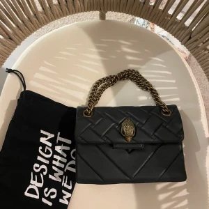 Svart quiltad handväska Kurt Geiger - Snygg svart quiltad handväska från Kurt Geiger med guldfärgad kedja som handtag och coolt emblem framtill. Väskan är tillverkad i skinn och har en stilren, boxig form. Perfekt för dig som gillar statement-accessoarer och vill ha något som sticker ut.
