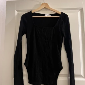 Svart ribbad långärmad topp NA-KD - Svart långärmad topp från NA-KD Reborn i storlek S. Toppen har en fyrkantig urringning och är ribbad i materialet vilket ger en snygg struktur. Nedre delen på toppen är även unik och snygg att para med ett par jeans!