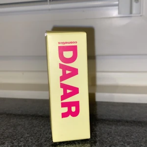 DAAR Cosmetics sminkprodukt - Säljer en sminkprodukt från DAAR Cosmetics i en snygg gul förpackning med rosa text. Perfekt för dig som gillar trendiga och färgglada märken. Exakt typ av produkt framgår inte av bilden, men DAAR är känt för sina innovativa sminkprodukter.