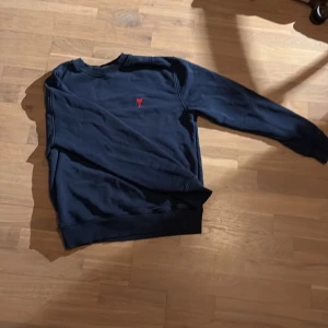Mörkblå sweatshirt från Ami - Snygg mörkblå sweatshirt från Ami med klassisk rund hals och långärmad passform. På bröstet finns ett litet broderat rött hjärta med bokstaven A. Tröjan är gjord i mjuk bomull och har ribbade muddar vid ärmslut och nederkant.
