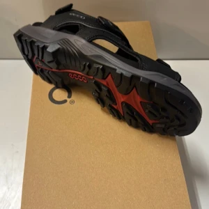 Svarta sandaler från ECCO med röd sula - Snygga svarta sandaler från ECCO med robust, mönstrad sula i svart och rött. Sandalerna har justerbara kardborreband och är tillverkade i slitstarkt syntetmaterial med detaljer i skinn. Perfekt för sommaren och utomhusaktiviteter.