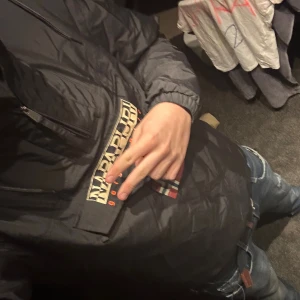 Svart vindjacka från Napapijri - Pris kan diskuteras. Svart vindjacka från Napapijri med huva och stor logga på bröstet samt norsk flagga. Jackan har lång ärm, elastiska muddar och en stor ficka framtill. Materialet är syntet och jackan är perfekt för blåsiga dagar. Snygg och stilren design med coola detaljer.