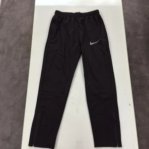 Svarta Nike byxor - Svarta Nike träningsbyxor med elastisk midja och silverfärgad logga på vänster lår. Byxorna har dragkedjor vid bensluten för enkel av- och påtagning. Materialet är lätt och syntetiskt, perfekt för sport och aktivitet. Storleken är M men är liten i storlek, ungefär S skriv vid funderingar. 