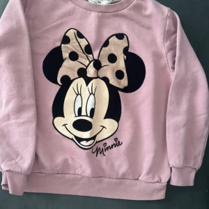 Rosa sweatshirt med Minnie Mouse - Söt rosa sweatshirt från H&M med stort tryck av Minnie Mouse på framsidan. Tröjan har en beige rosett med svarta prickar och ribbade muddar vid ärmslut och nederkant. Perfekt för dig som gillar Disney och vill ha en mysig och lekfull stil.