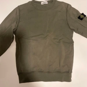Grön sweatshirt från Stone Island - Snygg grön sweatshirt från Stone Island med klassisk patch på vänster ärm. Tröjan har rund hals och långa ärmar, tillverkad i mjuk bomull. Perfekt för dig som gillar streetwear och vill ha en clean look med ikonisk detalj. Tröjan är för liten för mig och säljs därför. 