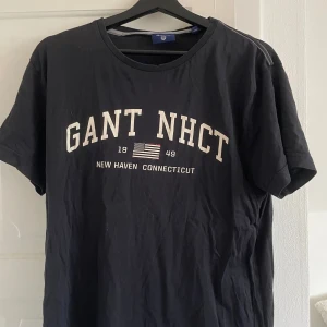 Svart GANT t-shirt med tryck - Snygg svart t-shirt från GANT med stort vitt tryck på bröstet där det står 'GANT NHCT' och en amerikansk flagga. Rund hals och klassisk passform, perfekt för dig som gillar college-stil. Materialet är mjuk bomull.