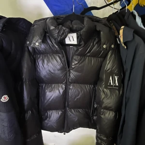 Svart pufferjacka från Armani Exchange - Säljer en svart pufferjacka från Armani Exchange i storlek S. Jackan har en glansig finish, huva med tryckknappar och dragkedja framtill. Stort AX-märke på ärmen och ribbade muddar vid ärmslut. Perfekt för kalla dagar när du vill ha både stil och värme.