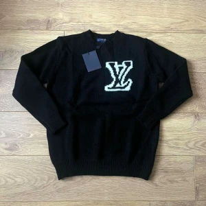 Tröja Louis Vuitton - Svart stickad tröja från Louis Vuitton med stor vit LV-logga på bröstet. Rund hals och långa ärmar med ribbade muddar. Perfekt för dig som gillar exklusiva märken och vill ha en stilren look.