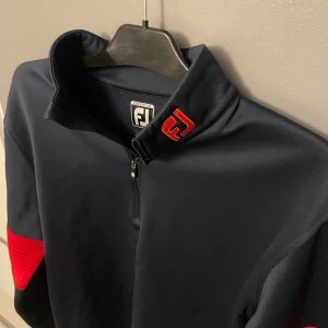 Blå footjoy half zip  - Snygg blå långärmad tröja från FootJoy med röd ribbad detalj längs ärmarna och ryggen. Tröjan har en hög krage med rött FJ-logo broderat och praktisk half zip framtill. Perfekt för dig som gillar sportig och modern stil. Tröjan är i sport material