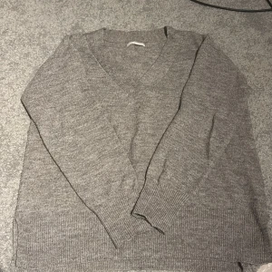 Grå stickad v-ringad tröja Ellos - Mysig grå stickad tröja från Ellos med v-ringad hals och långa ärmar. Tröjan har ribbade muddar vid ärmslut och nederkant, vilket ger en snygg och enkel look. Perfekt för att hålla värmen under kyliga dagar.