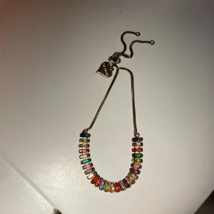 Färgglatt armband med stenar - Snyggt armband i guldton med rektangulära stenar i regnbågens alla färger. Justerbar kedja med hjärtberlock. Perfekt för dig som vill ha en färgklick på handleden och gillar unika smycken.