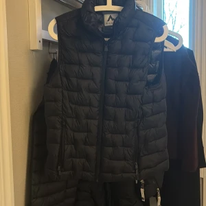 Svart dunväst från McKinley - Svart quiltad dunväst från McKinley med hög krage och dragkedja framtill. 