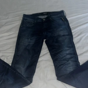 Replay Anbass jeans blå storlek 28 - Snygga Replay Anbass jeans i mörkblå tvätt med klassisk femficksdesign och diskreta slitningar. Modellen har normal passform och raka ben, med detaljerade sömmar och metallknapp framtill. Perfekta för dig som gillar stilrena och tidlösa jeans. Scick 9/10 då knappt använda. Priset är ej hugget i sten