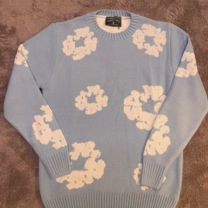 Denim teirs stickad - Säljer en ljusblå stickad tröja från Denim Teirs med vita blommiga mönster över hela tröjan. Rund halsringning och långa ärmar med ribbade muddar. Perfekt för dig som gillar mjuka material.