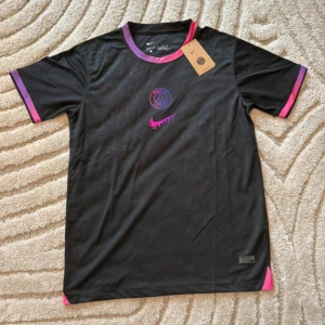 Svart PSG x Nike fotbollströja - Säljer en svart PSG fotbollströja från Nike med lila och rosa detaljer på krage och ärmslut. Tröjan har korta ärmar och är tillverkad i ett lätt, ventilerande material. PSG-logga och Nike swoosh i lila och rosa på bröstet. Perfekt för dig som gillar snygga matchtröjor.