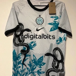Inter Milan fotbollströja Nike - Snygg Inter Milan fotbollströja från Nike med digitalbits-tryck på bröstet. Tröjan är vit med svarta och turkosa detaljer, har korta ärmar och coolt grafiskt mönster med ormar och blad. Rund hals med svart kant. Perfekt för dig som gillar unik design.