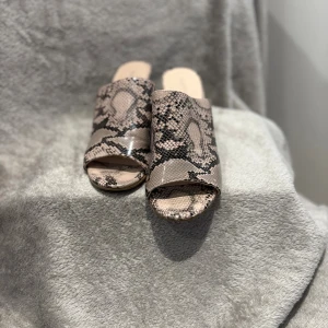 Beige sandaler med ormmönster och klack - Snygga sandaler från Pura Lopez med beige och svart ormmönster. Modellen har öppen tå, bred rem över foten och stabil klack i medelhöjd. Ytan är glansig och materialet känns som syntet med skinnstruktur. Perfekt för dig som gillar statement-skor.