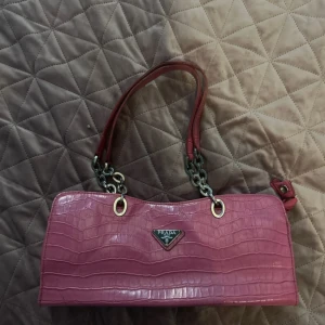 Rosa handväska i krokopräglat skinn Prada - Snygg rosa handväska från Prada i krokopräglat skinn med silverfärgade kedjedetaljer och dubbla handtag. Väskan har en rektangulär form och klassisk Prada-logga framtill. Perfekt för dig som vill sticka ut med en lyxig accessoar.  vet ej om den är äkta eller inte men finns ej att få tag i någonstans 