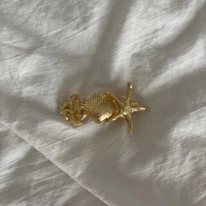 Guld hårspänne med snäckor och sjöstjärna - Unikt hårspänne i guldmetall med detaljerade snäckor och en sjöstjärna. Perfekt accessoar för dig som vill ha strandvibbar och coola detaljer i håret. Spännet har en klassisk klämma på baksidan som håller håret på plats.