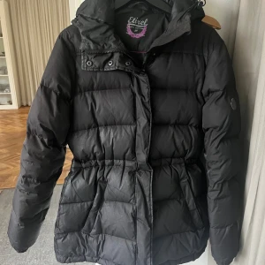 Svart pufferjacka från Etirel - Svart pufferjacka från Etirel med huva och snygga detaljer som emblem på ärmen. Jackan har dragkedja och knappar framtill, samt en markerad midja för schysst passform. Perfekt för kalla dagar och har en sportig vibe. Fodrad och riktigt skön att bära.