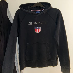  GANT Hoodie - Svart hoodie från GANT med broderad logga och amerikanskinspirerat sköldmärke på bröstet. Klassisk känguruficka framtill och huva. Perfekt för dig som gillar stilren och sportig look.