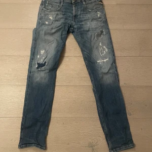 Replay anbass jeans med slitningar - Säljer ett par Replay Grover jeans i blå tvätt med coola slitningar och hål på benen. Klassisk femficksmodell med normal passform och raka ben. Jeansen har en snygg, lite blekt look och är tillverkade i jeansmaterial.