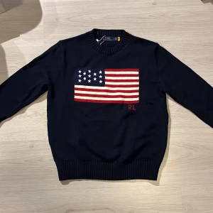 Mörkblå stickad tröja Polo Ralph Lauren - Mörkblå stickad tröja från Polo Ralph Lauren med amerikansk flagga broderad på bröstet och RL-detalj. Klassisk rund halsringning och ribbade muddar. Perfekt för dig som gillar preppy stil och vill sticka ut med en ikonisk detalj.