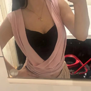 Rosa/svart vintage omlott ärmlös topp - Säljer en ljusrosa omlott topp med djup v-ringad urringning och draperad front. Toppen är ärmlös och har en snygg croppad passform som framhäver midjan. Underst finns en svart bandeau som är fastsytt på den rosa ger en cool kontrast. Perfekt för dig som gillar trendiga och unika plagg. Riktig vintage y2k