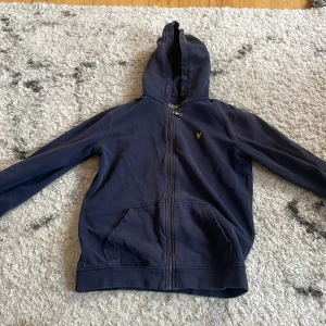 Marinblå hoodie från Lyle & Scott - Snygg marinblå hoodie från Lyle & Scott med dragkedja och huva. Tröjan har klassisk gul logga på bröstet och två stora fickor framtill. 