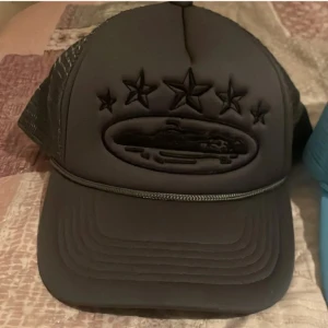 Svart keps med stjärnor och mesh - Snygg svart keps med mesh på sidorna och bak, samt broderade stjärnor och ett ovalt emblem framtill. Kepsen har böjd skärm och detaljerad brodyr som ger en cool look. Perfekt för dig som gillar streetstyle.