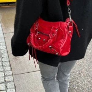 Liknande axelväska - Säljer en snygg röd axelväska med coola metalldetaljer och dragkedja framtill. Väskan har flätat handtag och flera fack, perfekt för att få plats med allt du behöver. Den har en urban vibe och sticker ut med sin starka färg.