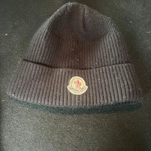 Svart ribbad Moncler mössa - Svart ribbstickad mössa från Moncler med klassisk logotyp framtill. Snygg och enkel design som passar perfekt när det är kallt ute. Mössan är tillverkad i mjukt material och har en uppvikt kant för extra stilpoäng.
