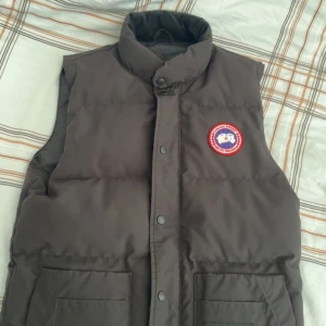 Svart dunväst från Canada Goose - Svart dunväst från Canada Goose med hög krage och tryckknappar framtill. Klassisk logga på bröstet och två stora fickor framtill. Västen är quiltad och har en clean, stilren look som funkar till många outfits.