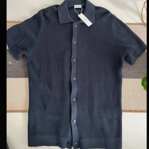 Marinblå stickad pikétröja John Henric - Snygg marinblå stickad pikétröja från John Henric med klassisk krage och knappar hela vägen framtill. Tröjan har korta ärmar och ribbad struktur som ger en cool och modern känsla. Perfekt för dig som gillar stilrena och tidlösa plagg. Helt ny med tagg.