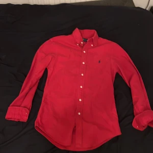 Röd skjorta från Ralph Lauren XS - Riktigt snygg röd skjorta från Ralph Lauren i slim fit-modell. Skjortan har klassisk krage, knäppning framtill och svart broderad logga på bröstet. Tillverkad i mjuk bomull och har långa ärmar med manschett.