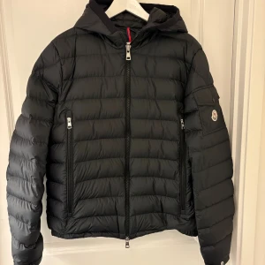 Svart Moncler dunjacka med huva - Svart dunjacka från Moncler med huva, quiltad design och flera dragkedjor. Jackan har Moncler-logga på ärmen och diskret branding i nacken. Fylld med dun och fjäder, perfekt för kalla dagar. Snygg, stilren och riktigt skön att bära. Jackan är i nyskick bara använd 2-3 gånger och är köpt för ungefär 1,5 månad sen. Jackan är köpt från CareOfCarl för 15.000kr kvitto finns även 