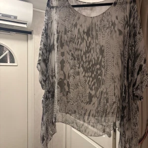 Grå transparent blus med mönster - Säljer en luftig och transparent grå-vit blus med abstrakt mönster. Blusen har en rund halsringning och är tillverkad i ett tunt, skönt material som faller snyggt. Perfekt för dig som gillar att sticka ut med coola detaljer och mönster.