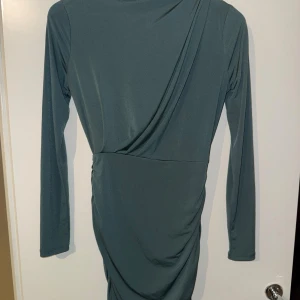 Mörkgrön omlottklänning Gina Tricot - Snygg mörkgrön kortklänning från Gina Tricot med lång ärm och draperad omlottdetalj framtill. Klänningen har en tight passform och är tillverkad i ett mjukt, stretchigt syntetmaterial som sitter skönt på kroppen.