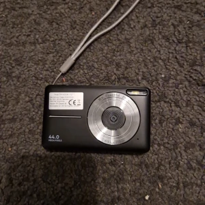 CMX-4032AF-11D Digitalkamera - Kompakt digitalkamera med 44 MP upplösning och 16x zoom. Levereras med laddare och skyddspåse. Kameran har en stor display och flera funktionsknappar. Skicket ser bra ut utan synligt slitage eller defekter.