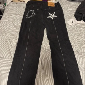 Svarta jeansbyxor från Corteiz - Svarta jeansbyxor från Corteiz med vita kontrastsömmar längs benen och broderade stjärn- och C-detaljer på bakfickorna. Klassisk femficksmodell med läderpatch och etikett. Snygg streetstil med rak passform. Storlek W32 endast testade. Ifall du behöver mått så skriv pm.