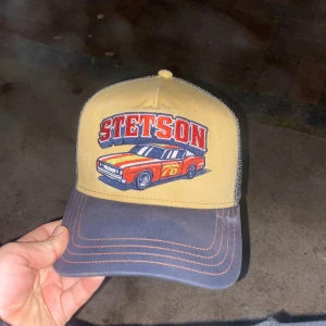 Stetson keps med biltryck - Cool keps från Stetson med broderad röd sportbil och STETSON-text framtill. Framsidan är beige, skärmen är grå med kontrastsömmar och baksidan har mesh i grått. Perfekt accessoar för dig som gillar bilar och streetstyle.