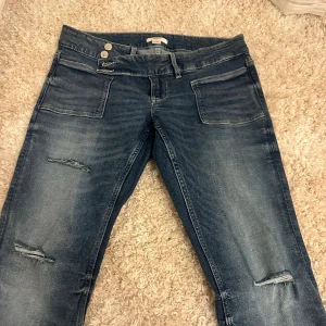 Blå bootcut jeans från Gina Tricot - Snygga blå jeans från Gina Tricot med bootcut och slitna detaljer på benen. Jeansen har två framfickor, dubbla silverknappar i midjan och är i väldigt bra skick. 