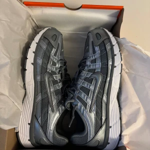 Nike p6000 - Nike Zoom Vomero 5 sneakers i metallicgrå och svart med vit sula. Ovandel i mesh och syntet med coola paneler och detaljer, samt klassisk Swoosh på sidan. Skorna har snörning och en chunky, sportig vibe som passar streetwear. Perfekta för dig som gillar techy och futuristisk stil.