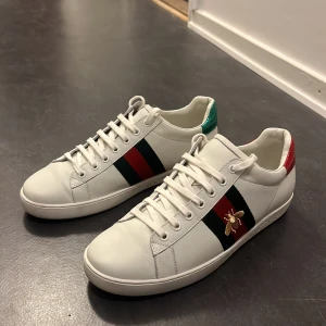 Gucci Ace sneakers med bi-detalj - Säljer ett par vita Gucci Ace sneakers i skinn med klassiska gröna och röda ränder på sidan samt broderad guldfärgad bi. Dom är knappt använda och i jätte fint skick. Äkta.  Storlek 43. 