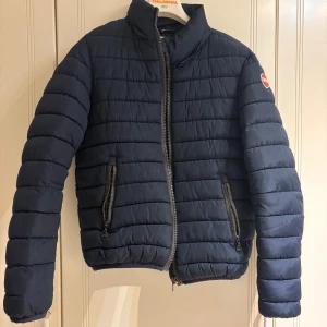 Mörkblå pufferjacka från Colmar - Säljer en mörkblå pufferjacka från Colmar Originals med snygga horisontella quiltade paneler och två praktiska fickor med dragkedja. Jackan har hög krage och Colmar-logga på ärmen. Perfekt för kyliga dagar och riktigt stilren. Priset kan diskuteras vid snabb affär