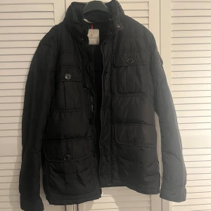 Svart Moncler dunjacka med fickor - Svart dunjacka från Moncler med fyra stora fickor framtill och en extra ficka på ärmen med Moncler-logga. Jackan har både dragkedja och knappar, hög krage och quiltad insida. Perfekt för kalla dagar och har en clean, urban look. storlek 3 M/L