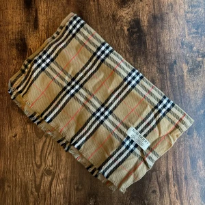 Burberry Halsduk - Elegant Burberry-halsduk i 100% lammull, tillverkad i England. Klassisk design med det välkända rutmönstret. Mycket bra skick, inga fläckar eller hål. Ett tidlöst plagg som lyfter varje outfit.
