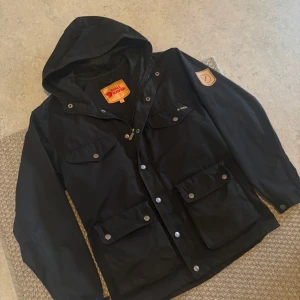 Svart Fjällräven parkajacka XS - Svart parkajacka från Fjällräven i G-1000 material, med huva och flera praktiska fickor med knappar och dragkedja. Klassisk Fjällräven-logga på ärmen och insidan. Perfekt för dig som gillar outdoor och stilren design. Passar S/M