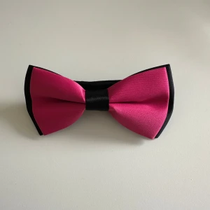 Svart och rosa fluga med justerbart band - Snygg fluga i svart och rosa med ett justerbart band och enkel spänne. Flugan har en klassisk form och är tillverkad i ett slätt syntetmaterial som ger en stilren look. Perfekt för att sticka ut med färg på festen eller balen.